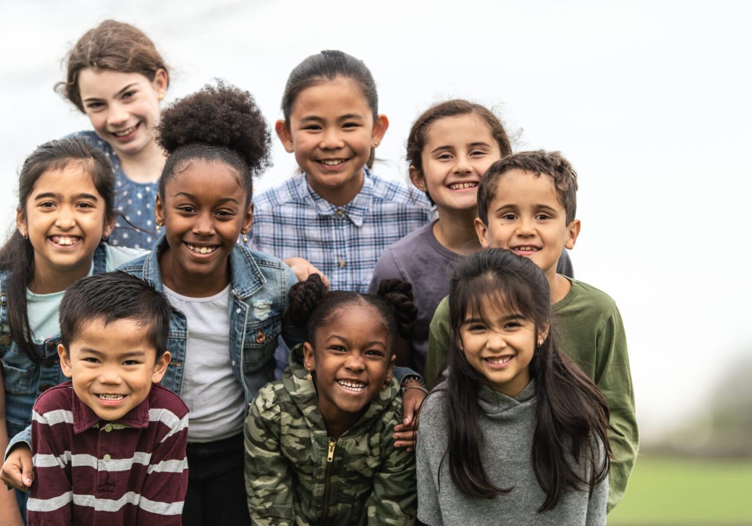 A group of multi-ethnic school aged children pause outside for a group portrait.  They are all leaning over and huddled in closely together, as they pose for the photo.  Each one is dressed casually and smiling as they enjoy the fresh air.