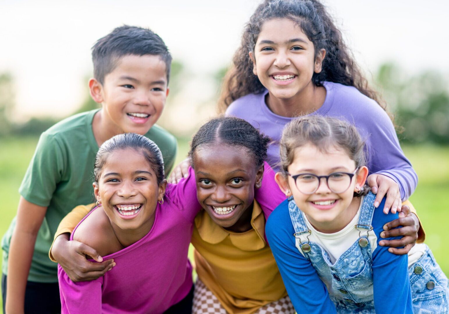 A small group of multi-ethnic school aged children huddle together outside as they pose for a portrait.  They are each dressed casually as they laugh and smile in the warm summer air.